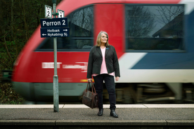 Ældre dame med demenssymbol på jakken venter på toget. 